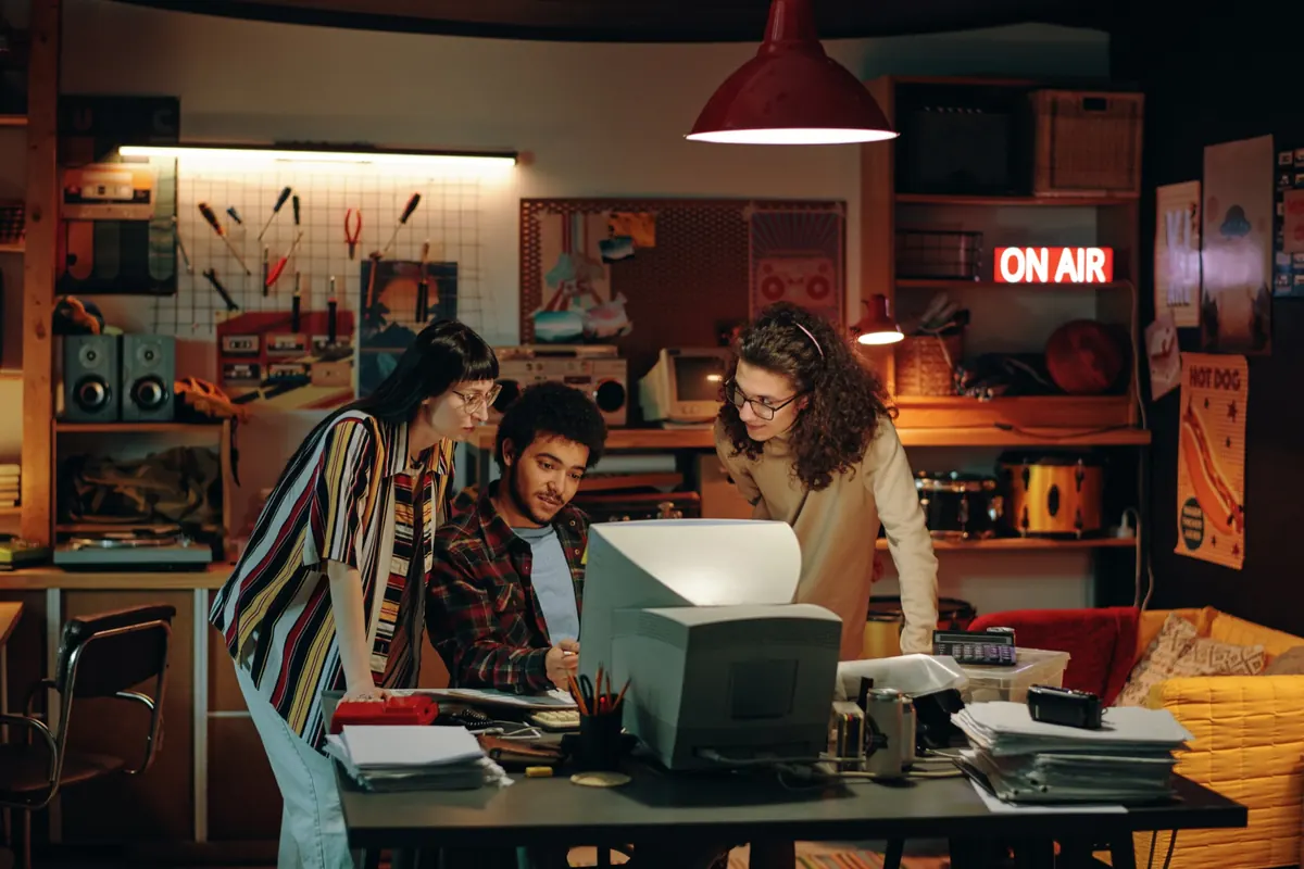 A small team gathered round a screen in a studio, an ON AIR sign lit behind them.