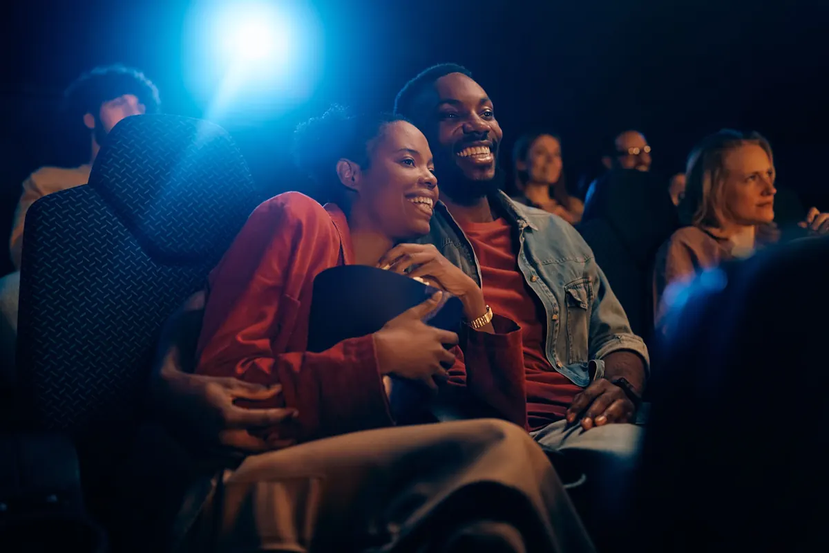 A couple laughing together in a cinema audience, popcorn between them.